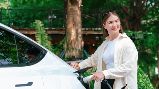 Young woman recharge her EV electric vehicle at green city park parking lot. Urban sustainability lifestyle for environmental friendly EV car with battery charging station. Expedient