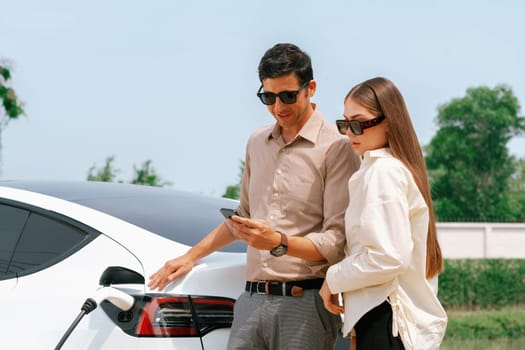 Young couple use smartphone to pay for electricity at public EV car charging station green city park. Modern environmental and sustainable urban lifestyle with EV vehicle. Expedient