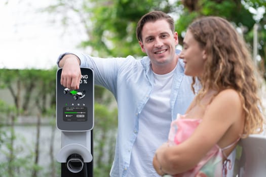 Happy and lovely couple with eco-friendly conscious recharging electric vehicle from EV charging station. EV car technology utilized as alternative transportation for future sustainability. Synchronos