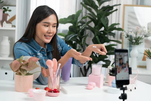 Young beautiful Asian girl reviewing moisturizing body lotion cream product to make glow skin testing on arm on social media online live recording smartphone with bestseller at modern room. Stratagem.