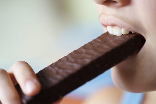 Closeup of crop anonymous teenage girl eating delicious chocolate protein bar at home