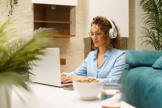 Positive curly haired female freelancer, in smart casual shirt and eyeglasses with headphones sitting at table while typing on laptop and working on remote project at home
