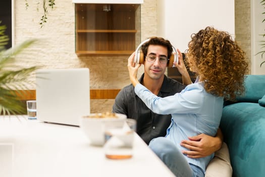 Pensive husband enjoying music over headphones, while smiling and holding curly haired wife and listening to music in wireless headphones in living room with laptop and cozy chair