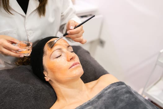 From above of crop anonymous cosmetician applying moisturizing product on skin of tranquil lady during beauty procedure in salon