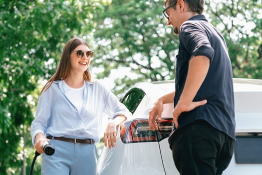 Lovely young couple wearing sun glasses recharging battery for electric car during road trip travel EV car in natural forest or national park. Eco friendly travel during vacation and holiday. Exalt