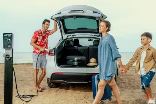 Family vacation trip traveling by the beach with electric car, lovely family taking luggage out while charging EV car battery with clean energy. Alternative family travel by eco-friendly car.Perpetual