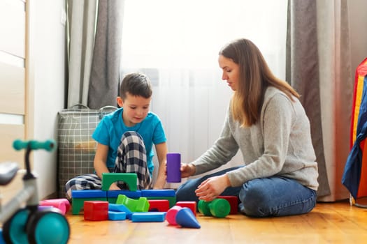 Young mother and her son playing together with colorful blocks, sitting on the floor at home