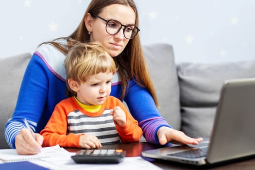 Young woman working at home with a laptop with a child on her lap. Home office with kids. Mother is working from home