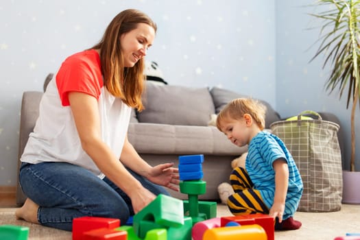 Cute little kid and child development specialist attractive young woman playing together with colorful blocks, sitting on the floor.