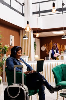 Smiling Young African American woman tourist wearing headphones using laptop while waiting for hotel check-in. Female traveler sitting with luggage at reception lounge area using computer