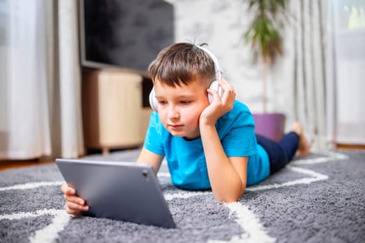 A child boy with tablet PC and headphones is lying on the floor watching cartoons, listening to music, playing games, communicates