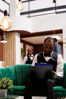 Computer and hotel industry. Black guy bellboy sitting with laptop in lobby entering data filling online document. African American man concierge making entry in daily luggage movement register