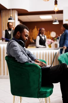 Focused black guy freelancer sitting in hotel lobby with laptop working remotely while traveling. Young African American man traveler using computer during business trip, waiting for check-in