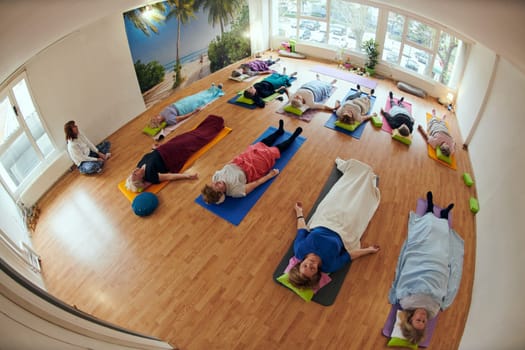 A group of senior women engage in various yoga exercises, including neck, back, and leg stretches, under the guidance of a trainer in a sunlit space, promoting well-being and harmony.