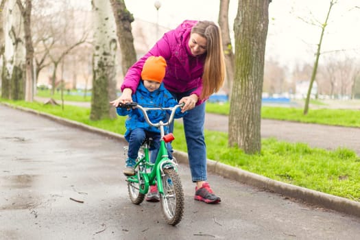 Mother teaching son to ride bicycle. Young mom teaching son to ride bike first time in park. Happy kids on bikes. The best moments together. Childhood bike concept