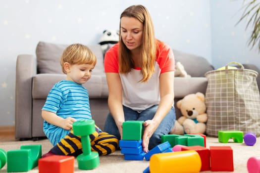 Cute little kid and child development specialist attractive young woman playing together with colorful blocks, sitting on the floor.