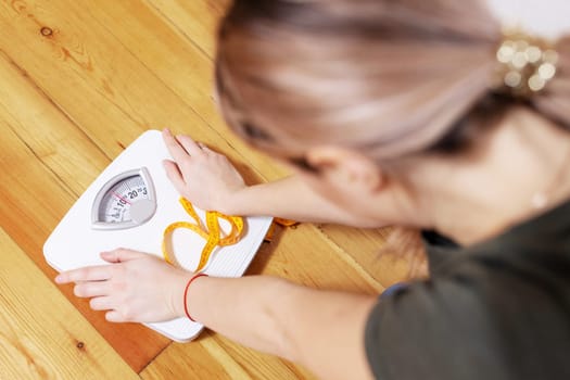 Depressed, frustrated and sad woman with a measuring tape looks at a white scale on a wooden floor