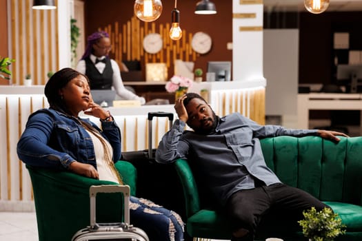 Tired African American man and woman having jet lag symptoms, resting with luggage in hotel lobby. Exhausted tourists feeling unwell after long-distance travel, waiting for check-in procedure