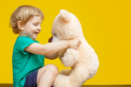 Little boy hugging teddy bear toy on yellow background.