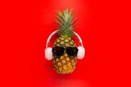 Ripe pineapple with sunglasses and headphones on red background. Copy space