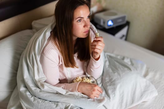 Young woman is watching projector, TV, movies and eating popcorn in the bed at home