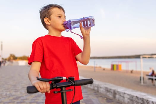A boy drinks water from plastic bottle, standing with two-wheeled scooter. Child playing outdoors with scooters. Outdoor activities.