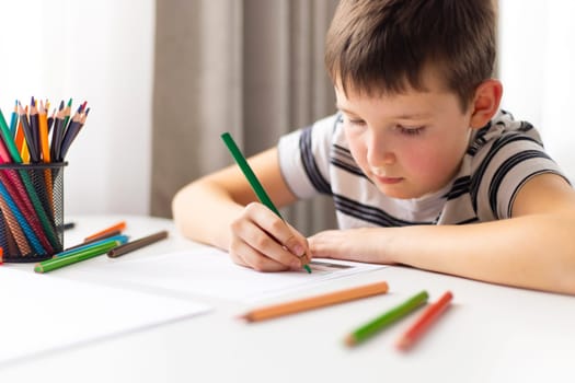 A child boy draws on white paper with colored pencils while sitting at a table.