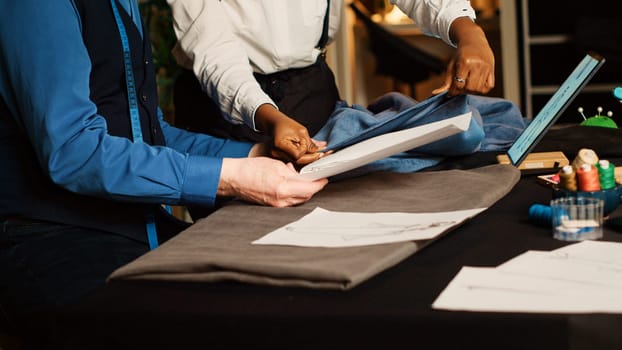 Old craftsman and trainee working in atelier, checking sketches of clothes to manufacture fashion design. Beginner learning the art of handcraft from experienced tailor. Handheld shot.