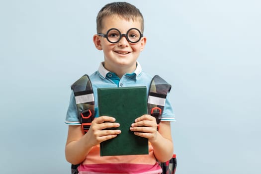 Elementary school nerd boy in eyeglasses with backpack holding book and looking at camera against blue background