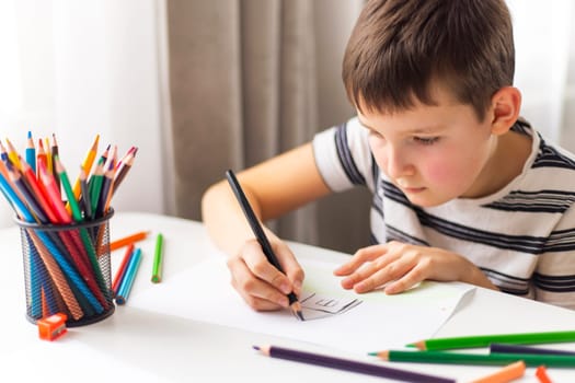 A child boy draws on white paper with colored pencils while sitting at a table.