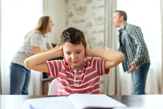 A sad child covers his ears with his hands during an argument between his parents. Family conflicts or divorce impact on on child development
