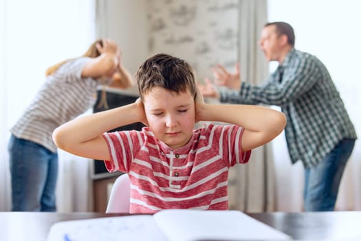 A sad child covers his ears with his hands during an argument between his parents. Family conflicts or divorce impact on on child development