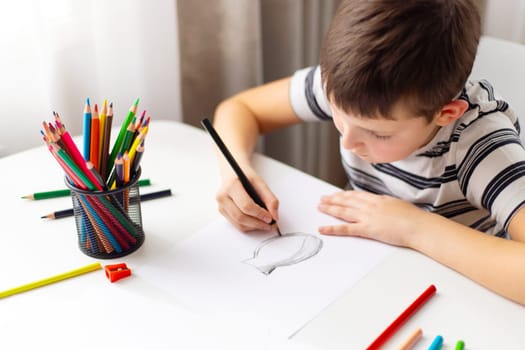 A child boy draws on white paper with colored pencils while sitting at a table.