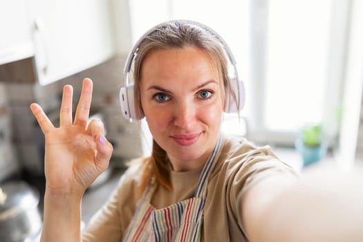 Young housewife in apron and earphones is talking on the phone and gestures ok in cozy kitchen