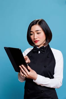 Restaurant young asian woman employee wearing uniform tapping on digital tablet. Cafeteria attractive waitress answering customer online message on portable gadget in studio