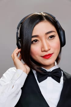 Beautiful asian waitress enjoying song playlist in headphones closeup. Young attractive woman receptionist wearing black and white uniform listening to music in earphones