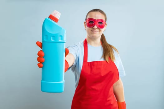 Young woman in red mask, rubber gloves and apron holding detergent bottle on blue background. Advertising detergent