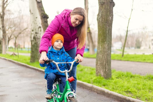 Mother teaching son to ride bicycle. Young mom teaching son to ride bike first time in park. Happy kids on bikes. The best moments together. Childhood bike concept