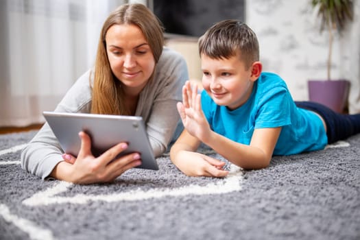 Happy mother and kid son greeting online looking at web camera using tablet pc for video call lying on carpet. Smiling mom and child having fun talking in video chat