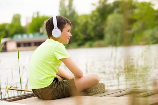 A boy with headphones listening to music at lakeside. Child sitting down by the lake listening to music