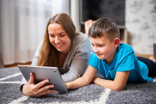 Happy loving family. Young mother and her son using tablet pc lying on carpet. Funny mom and lovely child are having fun at home