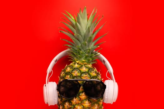 Ripe pineapple with sunglasses and headphones on red background. Copy space