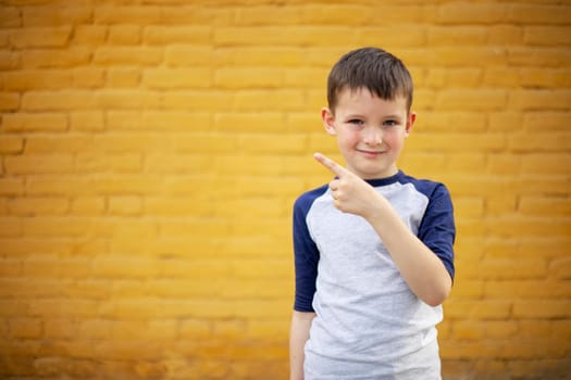 Smiling cute kid pointing with finger aside on yellow brick wall background.