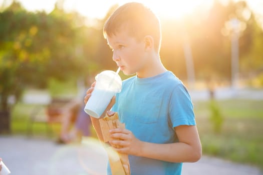 A child drinks a milkshake and eats a hotdog outdoors in a park.