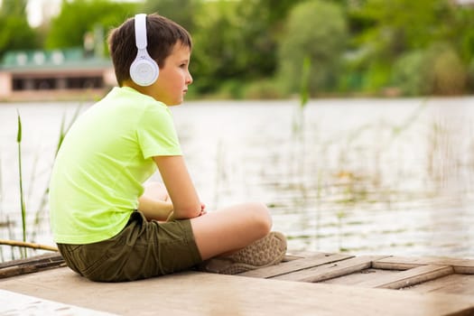 A boy with headphones listening to music at lakeside. Child sitting down by the lake listening to music