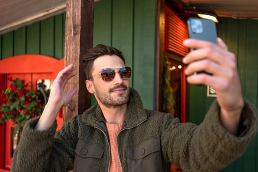 Handsome young man in trendy jacket and sunglasses taking selfie with smartphone. Male hipster making video chat via mobile phone outside wooden house.