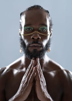 Portrait of serious African American gay man with blue eyeshadows and golden accessory on face. Shirtless adult transgender joining hands and looking at camera on blue background.