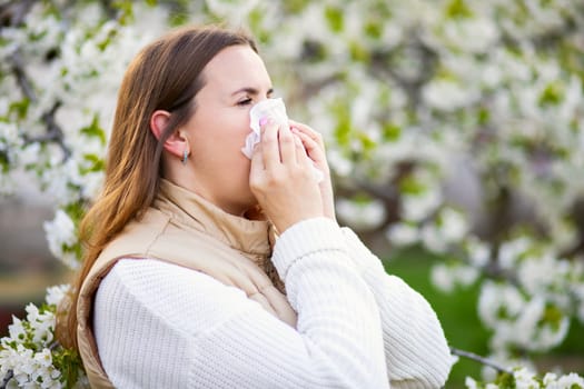 Sneezing woman suffering from seasonal allergy at spring with a nose wiper among the flowering trees in the park. Concept of spring allergies