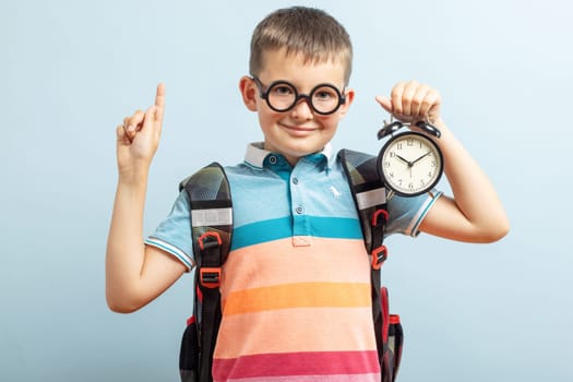 A cute school boy in glasses with an alarm clock against blue background. Hurry up. Time to go to school