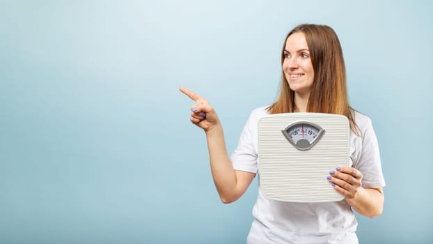 Young brunette woman with weighing scale pointing with finger to the side on blue background